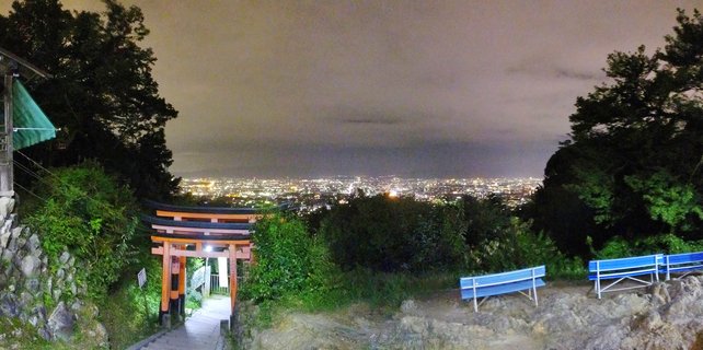 Night photograph showing an orange torii gate entrance with stone pathway, blue benches, and trees framing a panoramic view of illuminated Kyoto city lights in the distance under cloudy sky