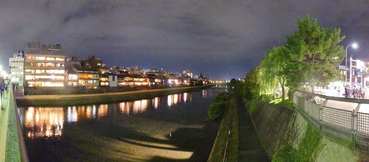 Night panoramic view of Kamogawa River in Kyoto showing illuminated traditional Japanese restaurants along the left bank with light reflections on water, trees and pedestrian walkway on right side