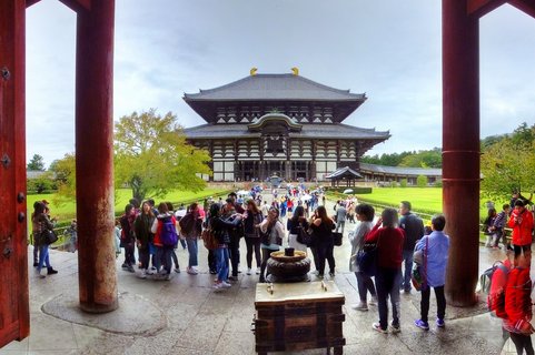 View through two large red wooden temple gate pillars showing Todai-ji Temple's Great Buddha Hall with tourists in the courtyard, green lawns, and autumn trees under cloudy sky