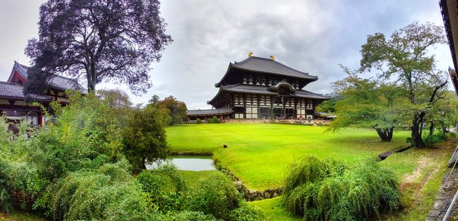 Wide view of Tōdai-ji Temple's Great Buddha Hall with traditional curved roof and golden ornaments, set against manicured green lawn and Japanese garden with pond and trees under cloudy sky