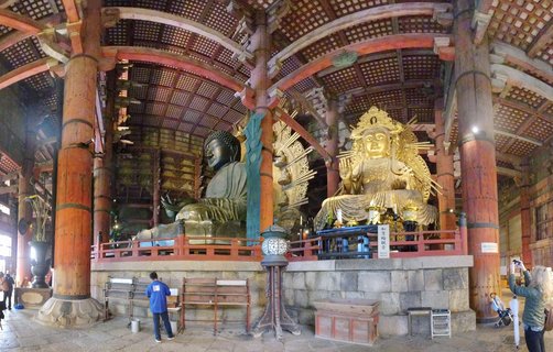 Interior view of Todai-ji Temple's Great Buddha Hall showing two massive Buddhist statues: the bronze Daibutsu on the left and golden Nyoirin Kannon on the right, with visitors below for scale