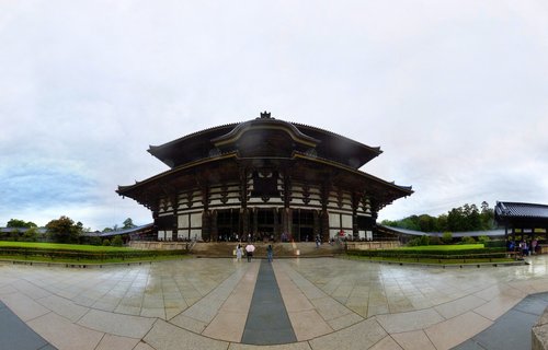 Large wooden Buddhist temple building with curved traditional Japanese roofs, stone courtyard with visitors walking toward entrance, overcast sky, Nara Japan