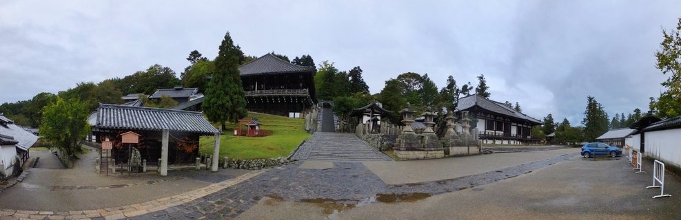 Traditional Japanese Buddhist temple complex with elevated wooden hall, stone lanterns, stone staircase, tiled roofs, and wet courtyard under cloudy sky