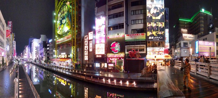 Night view of Dotonbori Canal in Osaka with colorful neon signs, the famous red Kani Doraku crab sign, building reflections in water, and pedestrians on the bridge walkway