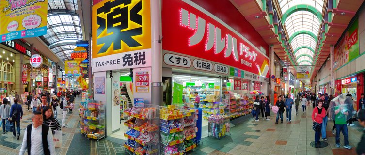 A covered shopping arcade in Japan with a red Tsuruha Drug store, colorful signs in Japanese, product displays, and pedestrians walking on tiled flooring under an arched glass ceiling
