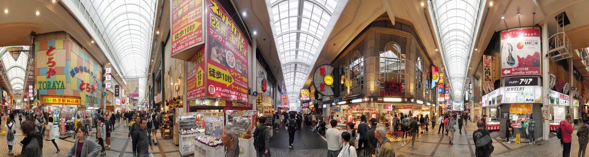 A wide covered shopping street in Japan with glass roof, multiple floors of shops with colorful Japanese signs and banners, and many people walking on the ground level