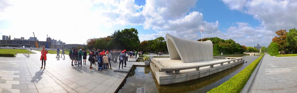 Wide view of the Hiroshima Peace Memorial Cenotaph with visitors walking on stone pathways, surrounded by water features, trees, and buildings under a blue sky with white clouds