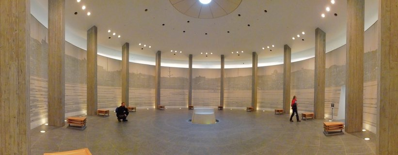 Interior view of circular memorial hall with concrete pillars, panoramic wall mural showing historical cityscape, wooden benches, and visitors walking through the space
