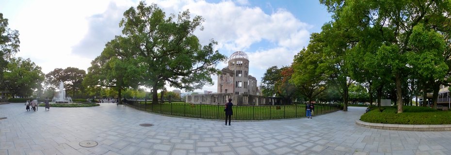 The Atomic Bomb Dome structure with its exposed steel framework dome stands in Hiroshima Peace Memorial Park surrounded by green trees, paved walkways, and visitors under a partly cloudy sky.