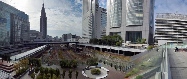 Wide urban cityscape showing multiple railway tracks running through central Tokyo with modern skyscrapers and the distinctive NTT Docomo Yoyogi Building tower visible against a partly cloudy sky.