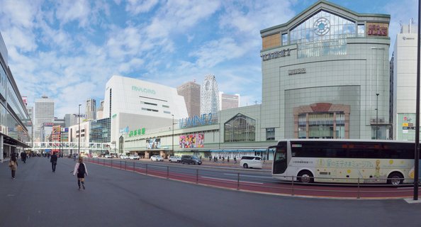 Wide street scene in Shinjuku Tokyo showing modern commercial buildings including Lumine 2 shopping center, with pedestrians walking and a tour bus on the road under blue sky with scattered clouds