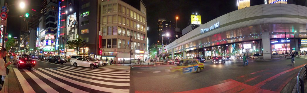 Nighttime panoramic street view of Roppongi district in Tokyo showing illuminated buildings with neon signs, vehicle traffic including taxis, pedestrian crosswalk, and people on sidewalks