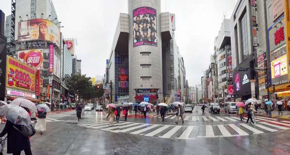 Aerial view of Shibuya Crossing in Tokyo with pedestrians holding umbrellas crossing wet streets, surrounded by buildings with large digital billboards and advertisements on an overcast day