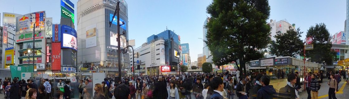 Aerial panoramic view of Shibuya Crossing in Tokyo showing dense crowds of pedestrians crossing a busy intersection surrounded by tall commercial buildings with Japanese signage and advertisements under a clear blue sky