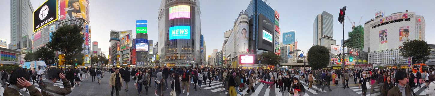 Aerial panoramic view of Shibuya Crossing in Tokyo showing large crowds of pedestrians crossing at a scramble intersection surrounded by tall buildings with digital billboards and commercial signs
