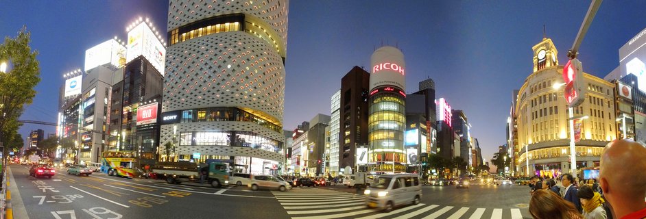 Panoramic view of Tokyo's Ginza district at twilight showing the Wako clock tower, modern buildings with neon signs including RICOH and UNIQLO, busy street intersection with vehicles and pedestrians under deep blue evening sky