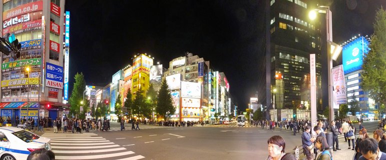 Night photograph of Shibuya Crossing in Tokyo showing crowds of pedestrians crossing a wide intersection surrounded by tall buildings covered in bright neon signs and billboards in Japanese text