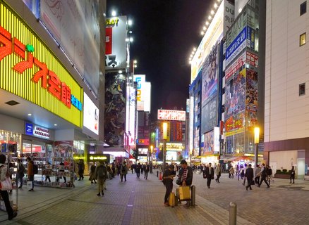 Night street scene in Akihabara Tokyo showing pedestrians walking on brick-paved street between buildings covered in bright neon signs and anime billboards