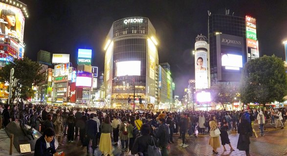 Night panoramic view of Shibuya Crossing in Tokyo showing crowds of pedestrians crossing the street surrounded by tall buildings with bright neon billboards and digital screens