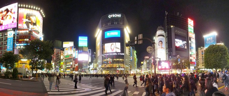 Night view of Shibuya Crossing in Tokyo showing crowds of pedestrians crossing the intersection surrounded by tall buildings with illuminated neon signs and large digital video screens including the QFRONT building