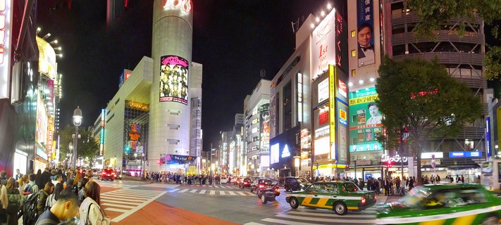 Night view of Shibuya Crossing in Tokyo showing crowds of pedestrians on crosswalks, green taxis, and buildings covered in bright neon signs and billboards including the cylindrical 109 building