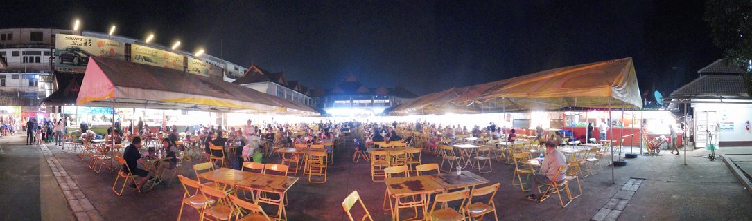 Wide panoramic photograph of an outdoor night market food court in Thailand showing numerous wooden tables and chairs arranged under large illuminated tents with people dining and walking around at night