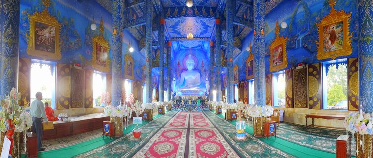 Interior view of a Buddhist temple with bright blue walls, golden decorative frames, a large white Buddha statue at the far end, and patterned red and green carpet leading toward it