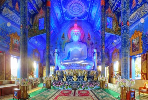 Large white Buddha statue in meditation pose inside an ornate blue Buddhist temple with gold pillars, floral offerings, and intricate ceiling murals