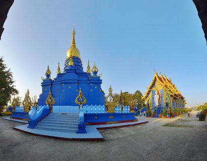 Vibrant blue Buddhist temple stupa with golden spire and ornate decorations, adjacent temple building with white Buddha statue, clear blue sky, Chiang Rai Thailand