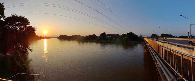 Wide river at sunset with golden sun reflection on water, concrete bridge with vehicles and motorcycles on right side, trees along left riverbank, power lines crossing sky