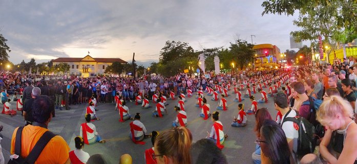 Traditional Thai cultural ceremony in Chiang Mai showing performers in white costumes with red sashes seated in rows on a street at twilight, surrounded by spectators, with colonial government building and Thai flag in background