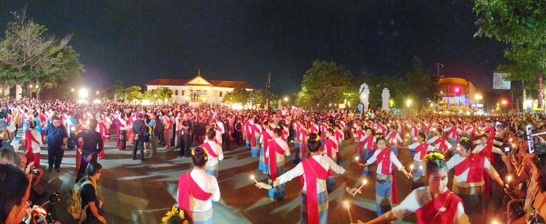 Large crowd at night festival in Thailand with women in traditional white and red clothing holding candles in procession, spectators watching and photographing, illuminated buildings in background