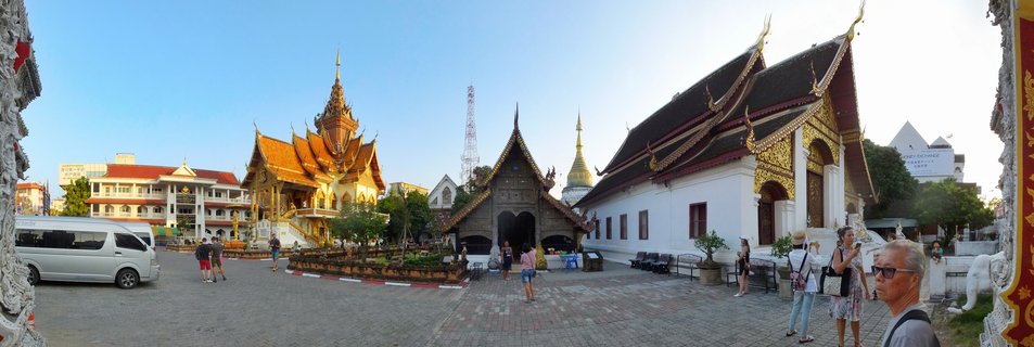 Panoramic view of a Thai Buddhist temple courtyard with multiple traditional buildings featuring ornate golden roofs, a golden stupa in the background, tourists walking on paved ground, and a white van parked on the left side under a clear blue sky