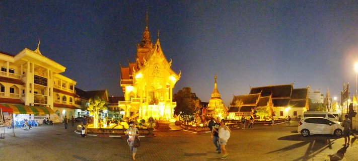 Panoramic photograph of an illuminated Thai Buddhist temple complex at dusk with golden buildings, a chedi stupa, and visitors walking through the courtyard under a deep blue sky