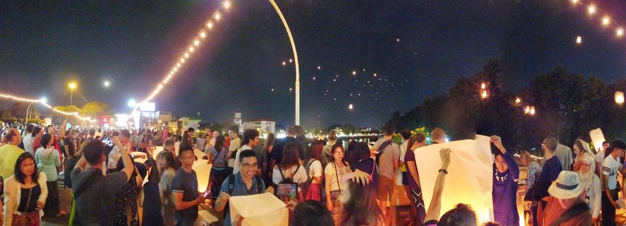 Large crowd of people at night releasing glowing paper sky lanterns during Yi Peng Festival in Chiang Mai, Thailand, with string lights overhead and multiple lanterns floating in the dark sky