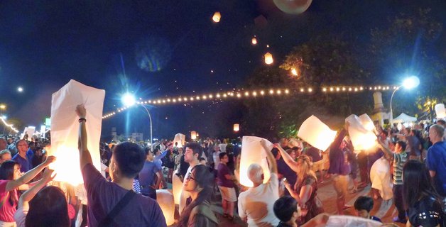 Large crowd of people releasing illuminated paper sky lanterns at night during Yi Peng Festival in Chiang Mai, Thailand, with multiple glowing lanterns floating upward against dark sky with string lights visible