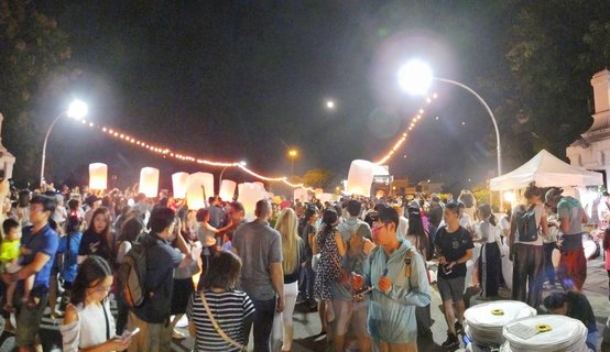 Large crowd of people at night releasing illuminated paper sky lanterns during Yi Peng Festival at Tha Phae Gate in Chiang Mai, Thailand with string lights overhead and full moon visible