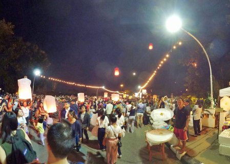 Large crowd of people releasing glowing paper sky lanterns into the night sky during Yi Peng Lantern Festival in Chiang Mai, Thailand, with string lights and street lamps illuminating the scene