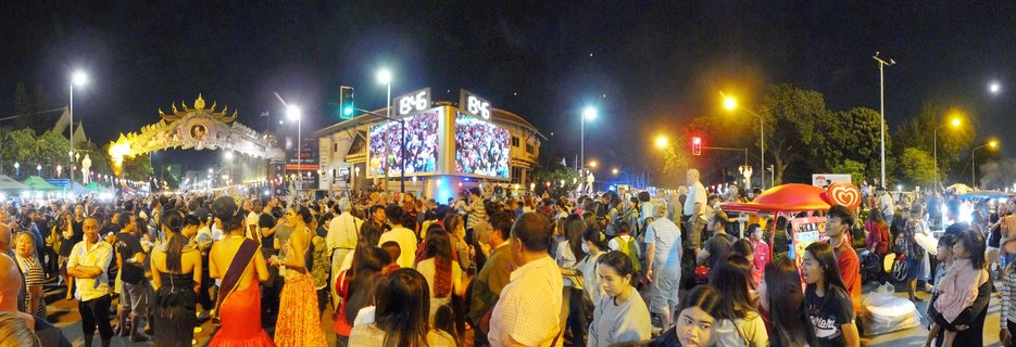Night photograph of a crowded street festival in Chiang Mai, Thailand showing an ornate golden Thai ceremonial gate with Lanna architecture, large LED screens, food vendor carts with red umbrellas, and hundreds of people filling the street under artificial lighting