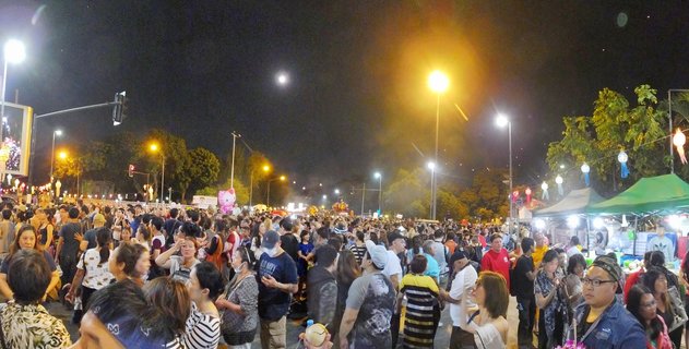 Large crowd of people gathered on a street at night in Chiang Mai, Thailand. Street lights illuminate the scene with market stalls and hanging lanterns visible on the right side. Full moon visible in the dark sky.