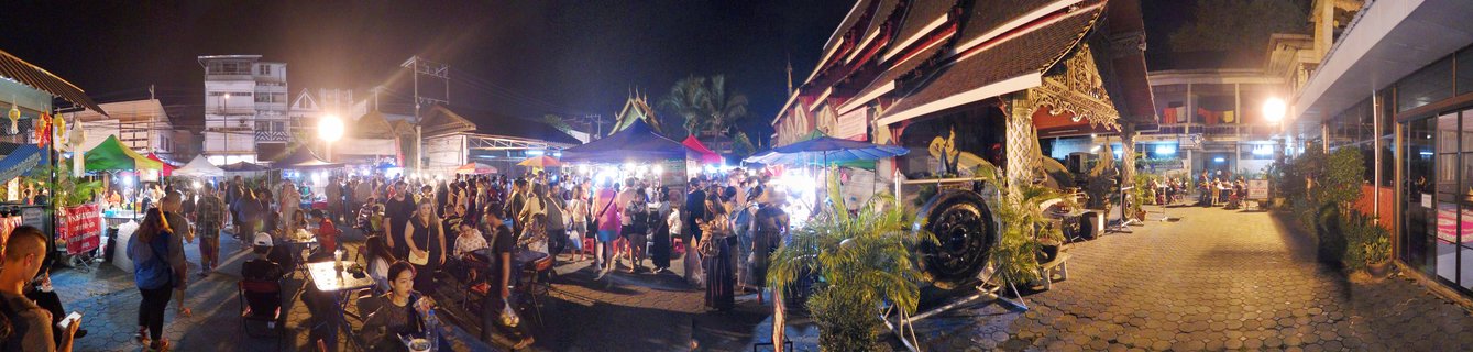 Wide panoramic view of a crowded night market street in Chiang Mai with people walking between vendor stalls under colorful tents, traditional Thai temple building with ornate roof on the right side, artificial street lighting throughout