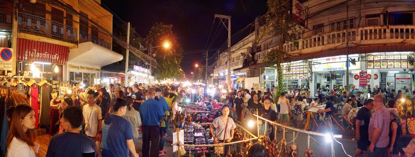 A crowded street market at night in Chiang Mai with people browsing vendor stalls, illuminated shop signs, and restaurants lining both sides of the pedestrian area