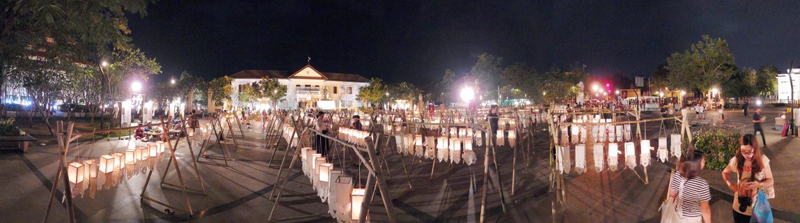 Night photograph showing multiple rows of illuminated white paper lanterns hanging from wooden A-frame structures in an outdoor plaza, with people walking among the displays and a white building visible in the background