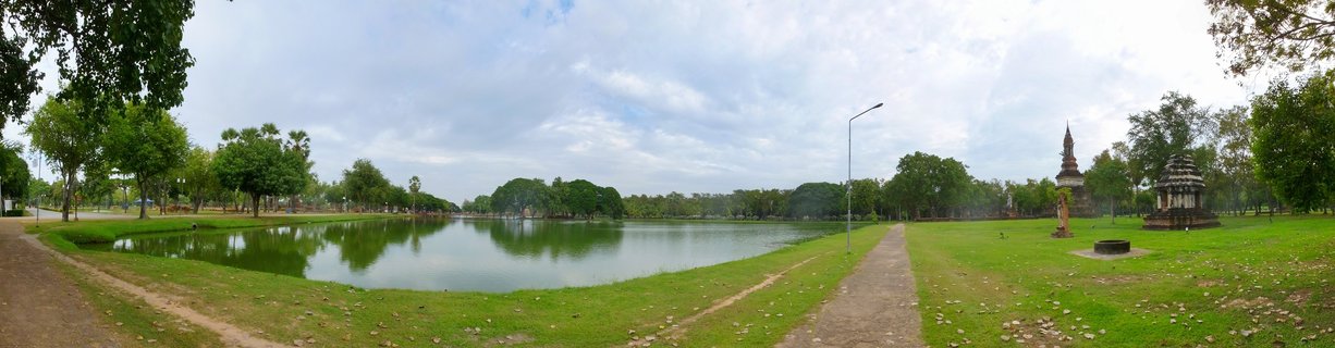 Panoramic view of Sukhothai Historical Park showing ancient Buddhist temple chedis and ruins beside a calm lake with green grass, trees, and cloudy sky
