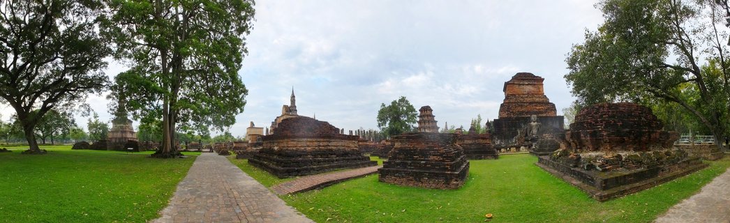 Panoramic view of ancient brick Buddhist temple ruins with multiple stupas and chedis surrounded by green lawns and large trees at Sukhothai Historical Park in Thailand