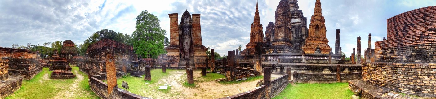 Panoramic view of ancient brick Buddhist temple ruins featuring multiple stupas, chedis, and a large standing Buddha statue at Sukhothai Historical Park in Thailand under cloudy sky