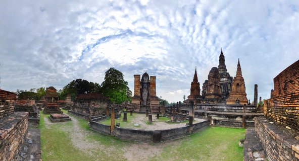 Panoramic view of ancient brick Buddhist temple ruins with multiple stupas and a standing Buddha statue at Sukhothai Historical Park in Thailand under cloudy sky