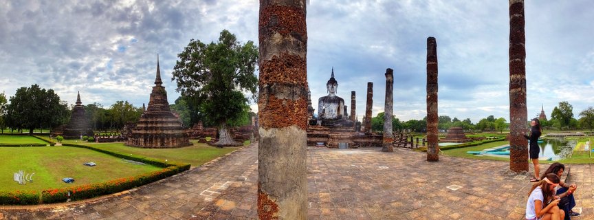 Large seated Buddha statue surrounded by ancient brick columns and multiple stupas at Sukhothai Historical Park in Thailand with tourists visiting the UNESCO World Heritage Site under cloudy sky
