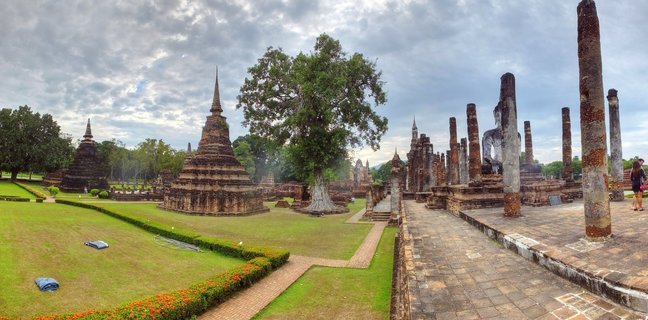 Panoramic view of ancient Buddhist temple ruins showing multiple brick stupas, stone columns, and Buddha statues at Sukhothai Historical Park in Thailand with green lawns and cloudy sky
