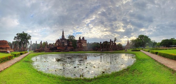 Ancient Buddhist temple ruins with brick stupas and spires reflected in a lotus pond surrounded by green lawns and pathways under cloudy sky at Sukhothai Historical Park, Thailand